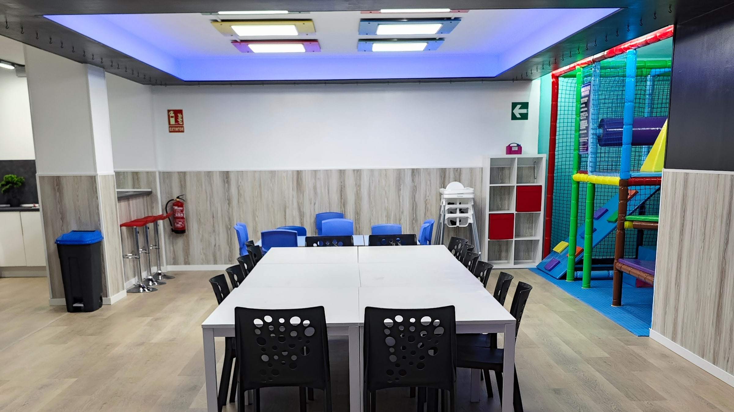 Zona de mesas con sillas para adultos y el parque infantil de bolas al fondo de la sala