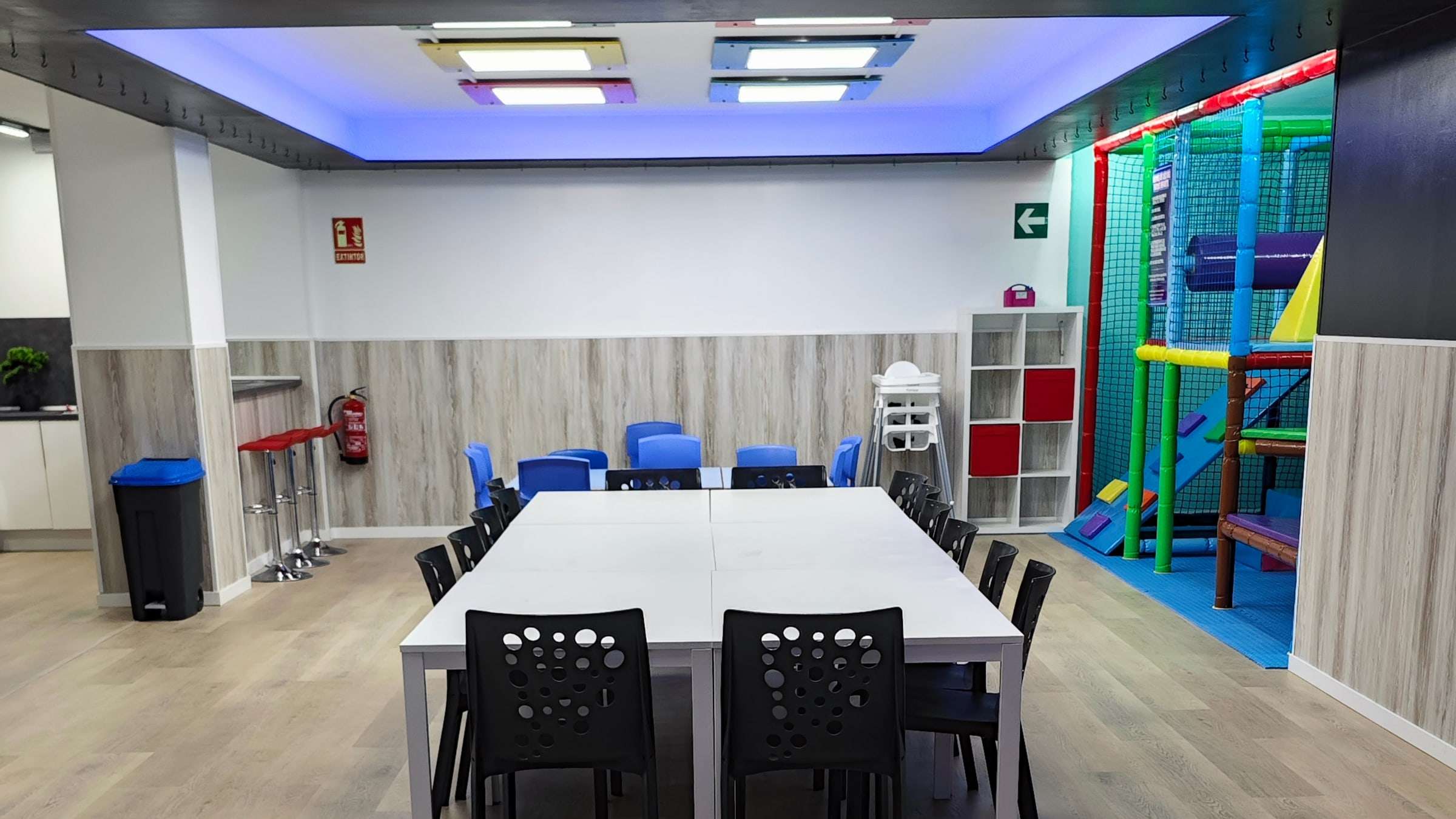 Zona de mesas con sillas para adultos y el parque infantil de bolas al fondo de la sala