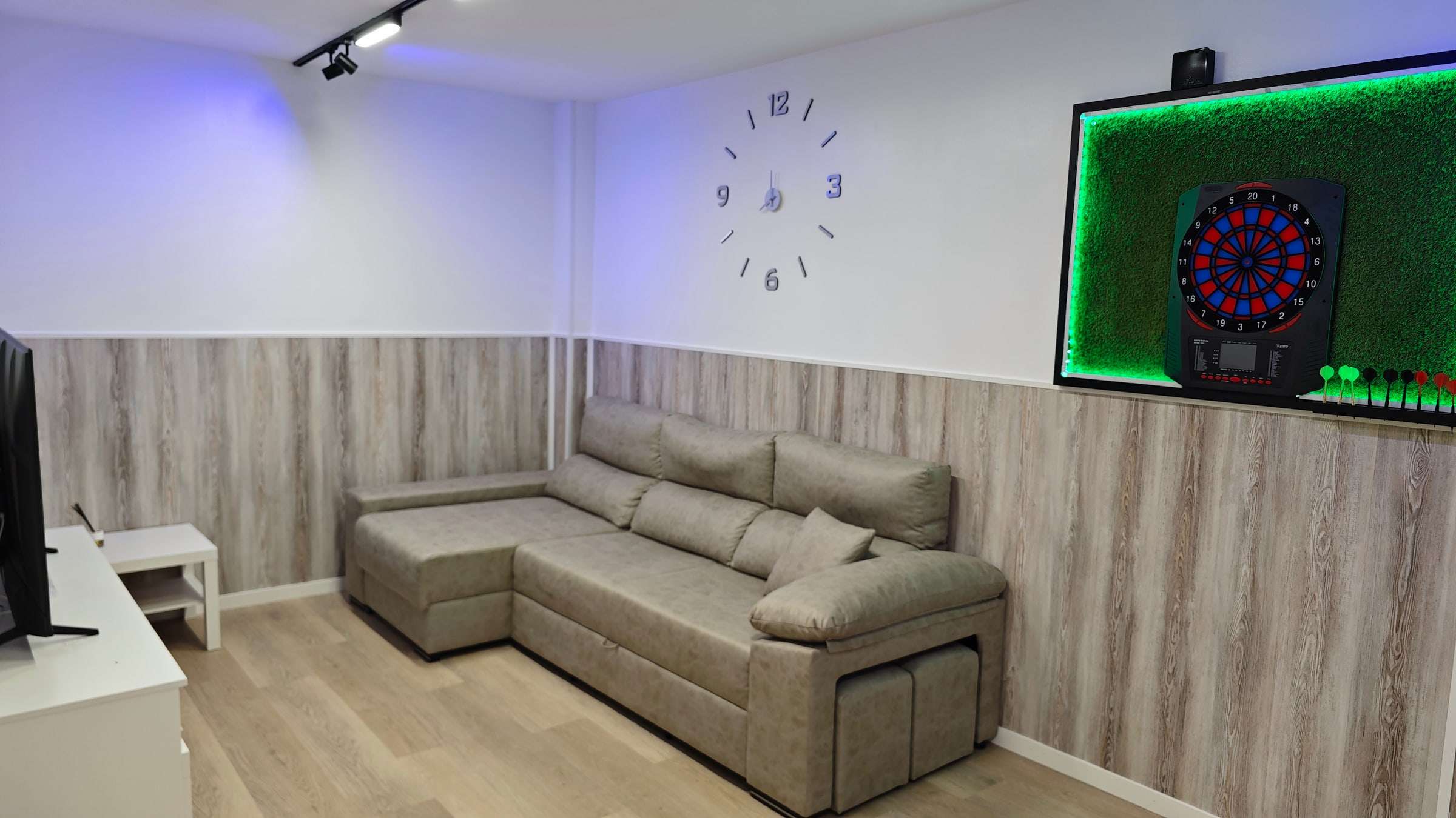 Zona relax con sofá cama gris y diana electrónica de dardos en la pared