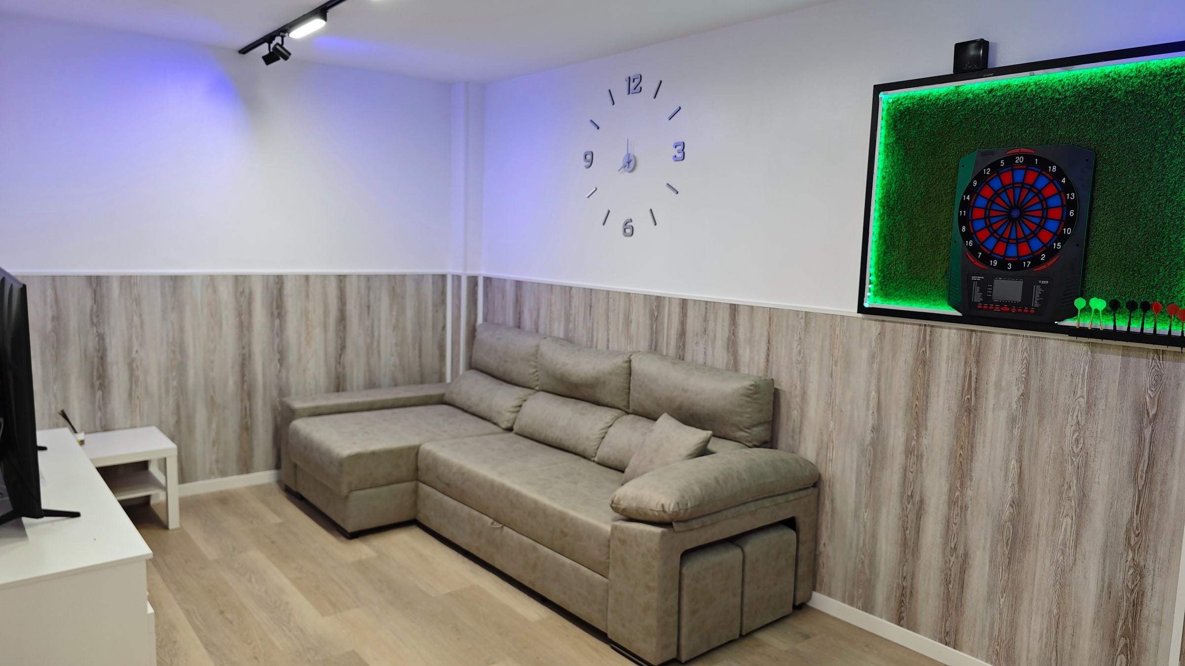 Zona relax con sofá cama gris y diana electrónica de dardos en la pared